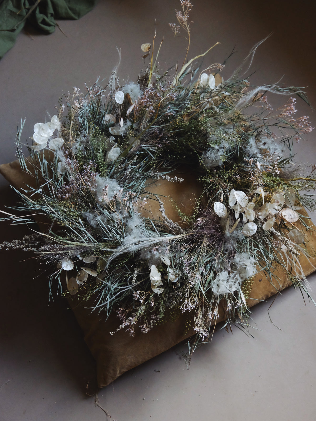 A dried floral wreath with white flowers, sage green foliage, and soft beige grasses, displayed on a brown cushion against a neutral background.