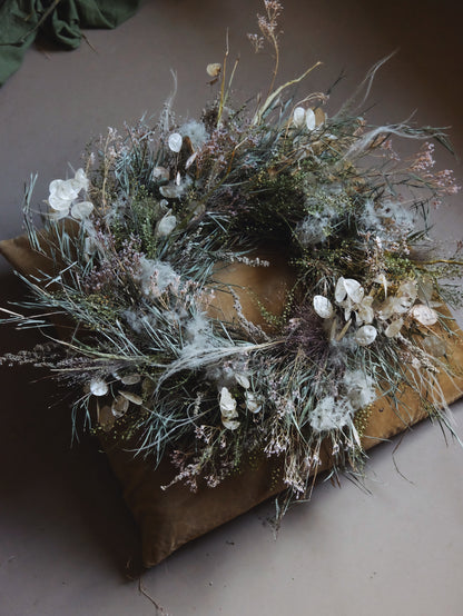 A dried floral wreath with white flowers, sage green foliage, and soft beige grasses, displayed on a brown cushion against a neutral background.