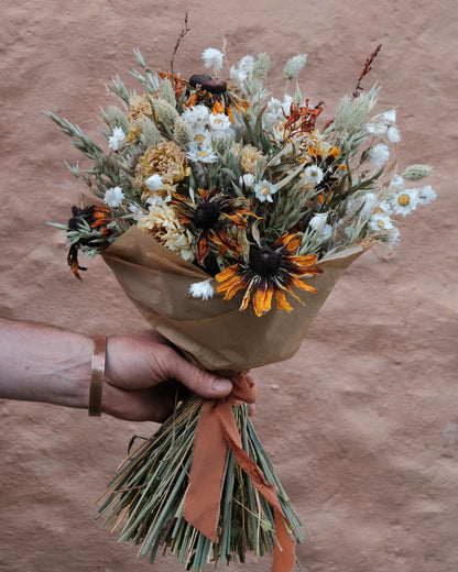 A hand holding a dried flower bouquet wrapped in brown kraft paper with an orange-brown ribbon, set against a textured terracotta wall.