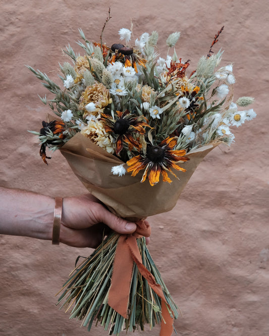 A hand holding a dried flower bouquet wrapped in brown kraft paper with an orange-brown ribbon, set against a textured terracotta wall.