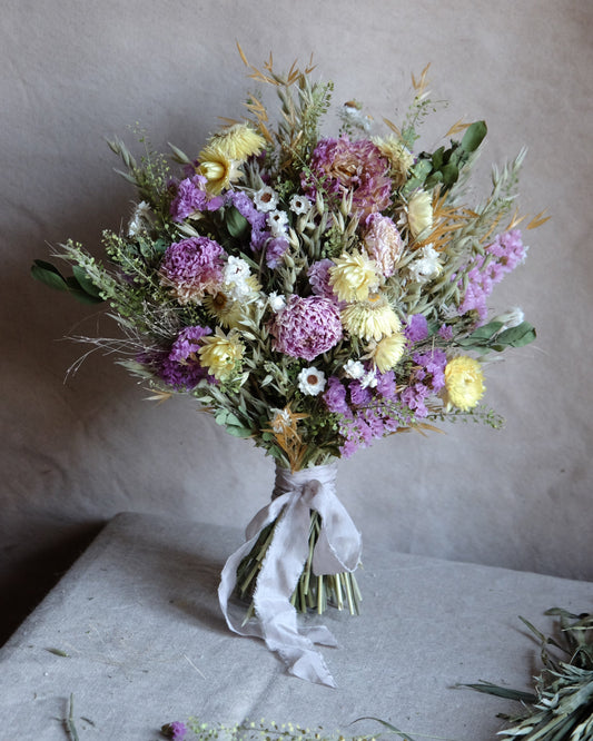 A rustic dried flower bouquet in purple, yellow, and white tones with green stems, tied with a white ribbon, displayed on a neutral gray surface against a textured beige background.