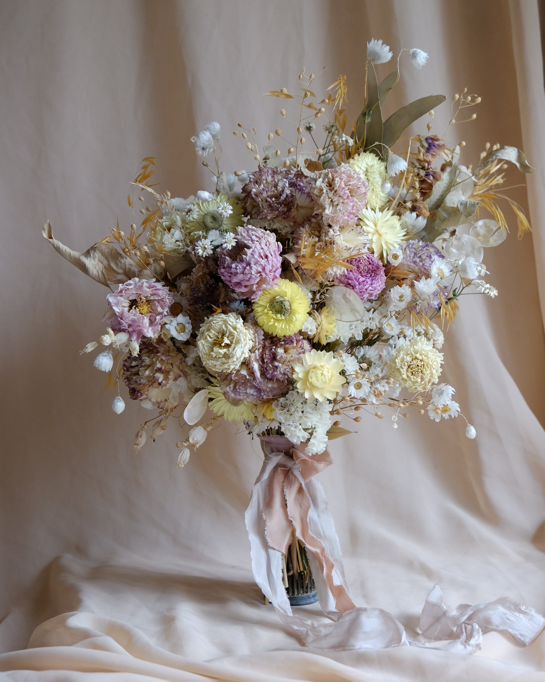 A dried flower bouquet in soft pastel tones, including cream, lavender, dusty pink, and muted yellow, tied with pale pink ribbons and set against a draped beige fabric backdrop.