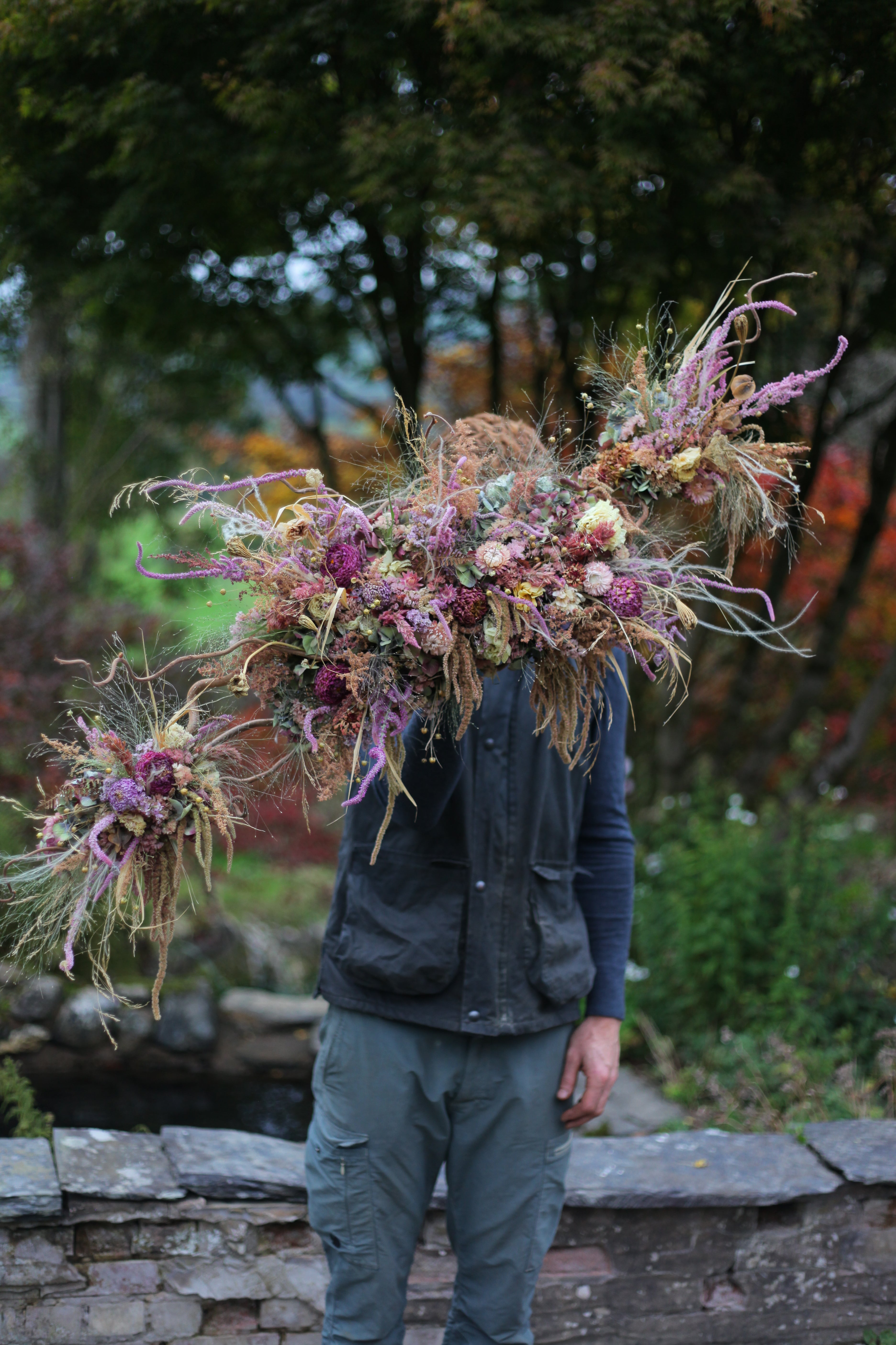 Person holding a large, intricate floral arrangement outdoors with stone wall and greenery in the background.