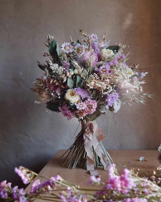 A dried flower bouquet with lavender, pink, cream, and green elements tied with a blush ribbon, displayed on a wooden surface against a textured brown wall.